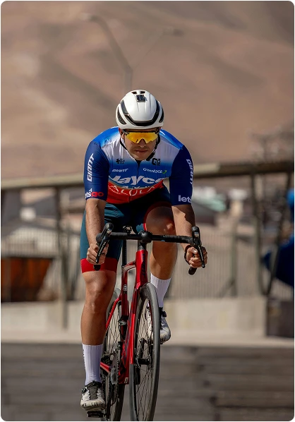 Un cycliste portant un casque blanc et une tenue bleue roule sur un vélo rouge par une journée ensoleillée, concentré et intense. Les collines désertiques forment un arrière-plan flou.