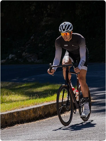 Un cycliste vêtu d'une tenue noire et blanche roule sur une route ensoleillée, concentré et déterminé. De la verdure est visible sur le bord de la route, projetant des ombres sur le chemin.