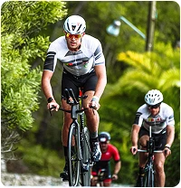 Des cyclistes portant des casques et des tenues assorties roulent sur une route entourée d'une végétation luxuriante. Le cycliste de tête fait preuve de détermination et de concentration.