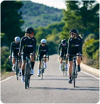 Un groupe de cyclistes vêtus de tenues noires roule sur une route panoramique entourée d'une végétation luxuriante. L'ambiance est énergique et concentrée sous un ciel clair.