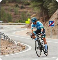 Un cycliste vêtu d'un maillot bleu et d'un casque descend une route de montagne sinueuse, concentré. En arrière-plan, on aperçoit des arbres et des panneaux de signalisation.