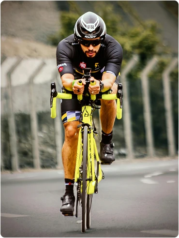 Un cycliste vêtu d'une tenue sombre et d'un casque roule sur une route avec un vélo jaune vif. L'arrière-plan est flou, avec une clôture et de la verdure, ce qui donne une impression de vitesse et de concentration.