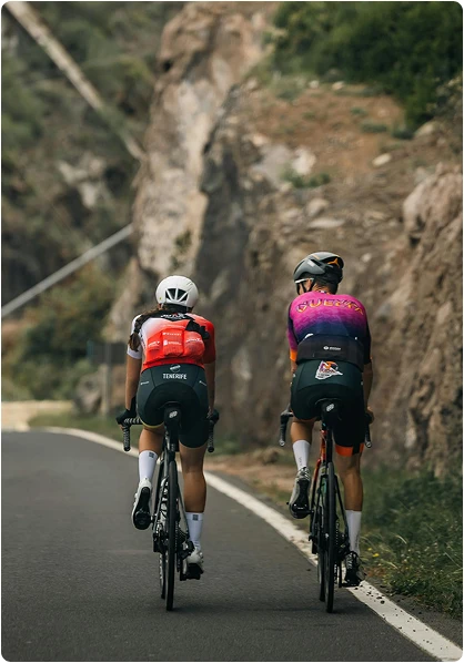 Deux cyclistes gravissent une route sinueuse bordée de falaises rocheuses et de verdure. Ils portent des maillots et des casques colorés, et respirent la concentration et la détermination.